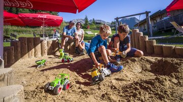 Zwei Kinder spielen mit Spielzeugtraktoren im Sandkasten. Im Hintergrund sitzen Erwachsene unter roten Sonnenschirmen.