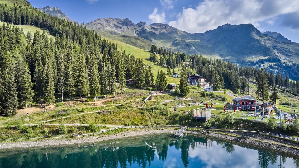 Eine malerische Berglandschaft mit grünen Wäldern und sanften Hügeln. Ein ruhiger See spiegelt die Umgebung wider.