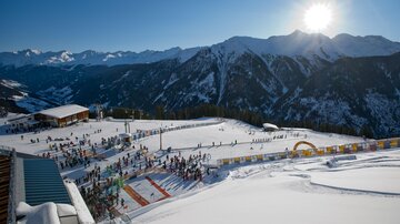 Eine winterliche Berglandschaft mit Skifahrern und sonnigem Himmel. Im Vordergrund sind schneebedeckte Pisten und eine belebte Skischule zu sehen.