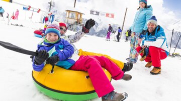 Eine Gruppe von Kindern und Erwachsenen hat Spaß im Schnee. Sie fahren auf Reifen und lachen in der winterlichen Landschaft.