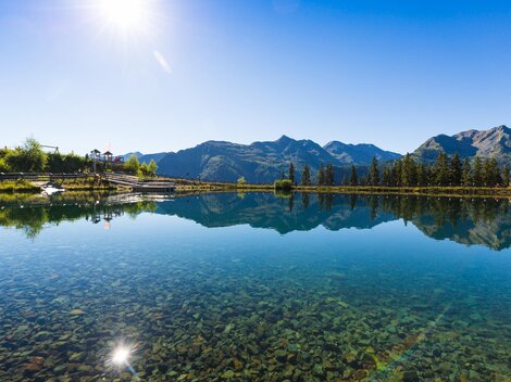 Ein ruhiger See spiegelt die Berge und den blauen Himmel wider. Die Umgebung ist grün und sonnig.