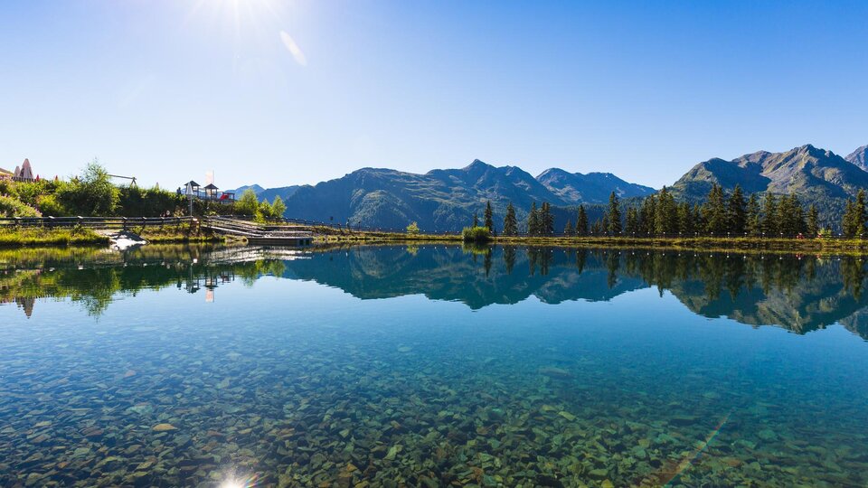 Ein ruhiger See spiegelt die Berge und den blauen Himmel wider. Die Umgebung ist grün und sonnig.