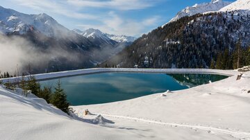 Ein klarer Bergsee umgeben von schneebedeckten Bergen. Der Himmel ist blau mit einigen Wolken.