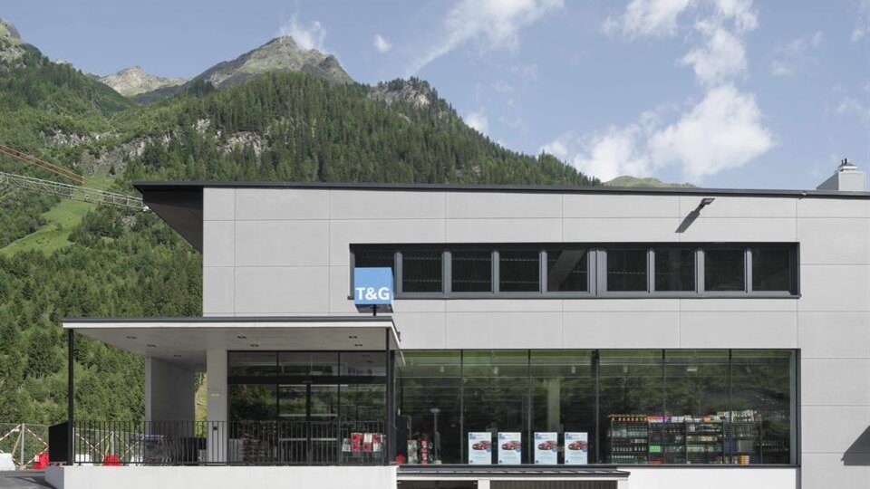 Ein modernes Gebäude mit großen Fenstern und einem Parkplatz davor. Im Hintergrund sind grüne Berge unter einem blauen Himmel zu sehen.