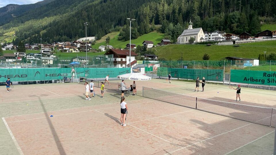 Ein Tennisplatz mit mehreren Spielern und einer bergigen Landschaft im Hintergrund. Die Umgebung ist grün mit typischer alpenländischer Architektur.