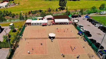 Eine Tennisanlage mit mehreren Spielfeldern und Spielern. Im Hintergrund sind Gebäude und Fahrzeuge zu sehen, umgeben von grüner Landschaft.