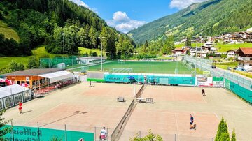 Ein schöner Tennisplatz in einer grünen Landschaft mit Bergen im Hintergrund. Einige Spieler sind aktiv, während andere auf der Bank sitzen.