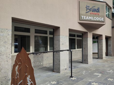 Ein modernes Gebäude mit einem Schild, das "Teamlodge" anzeigt. Vor dem Eingang steht ein weiteres Schild mit sportlichen Motiven.