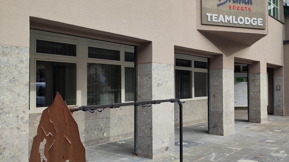 Ein modernes Gebäude mit einem Schild, das "Teamlodge" anzeigt. Vor dem Eingang steht ein weiteres Schild mit sportlichen Motiven.