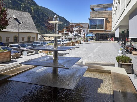 Ein moderner Platz mit einem Wasserfall und Sitzmöglichkeiten. Im Hintergrund sind Gebäude und Parkplätze in einer malerischen Umgebung zu sehen.