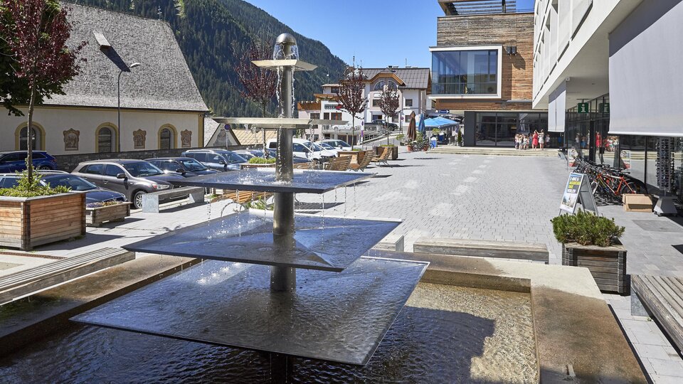Ein moderner Platz mit einem Wasserfall und Sitzmöglichkeiten. Im Hintergrund sind Gebäude und Parkplätze in einer malerischen Umgebung zu sehen.