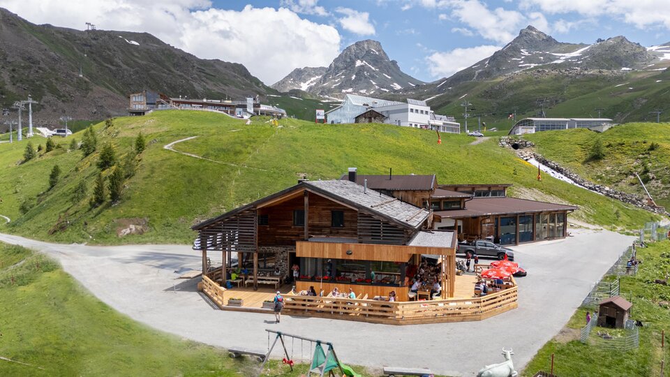Ein modernes Holzhaus in den Bergen, umgeben von grünen Wiesen und schroffen Gipfeln. Der Himmel ist blau mit einigen weißen Wolken.