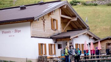 Eine gemütliche Alm mit Holzfassade und mehreren Tischen im Freien. Wandersleute stehen vor dem Eingang und genießen die Berglandschaft.