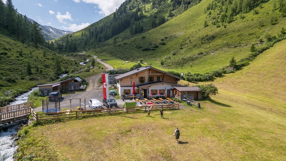Eine malerische Landschaft mit einem Gasthaus in den Bergen. Fluss und grüne Wiesen umgeben das Gebäude.