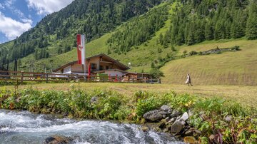 Eine malerische Almhütte umgeben von grünen Wiesen und hohen Bergen. Ein klarer Bach fließt im Vordergrund.