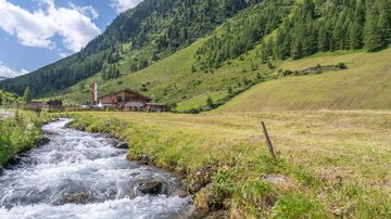Ein klarer Bach fließt durch eine grüne Wiese, umgeben von sanften Hügeln und bewaldeten Bergen. Im Hintergrund ist ein rustikales Haus sichtbar.