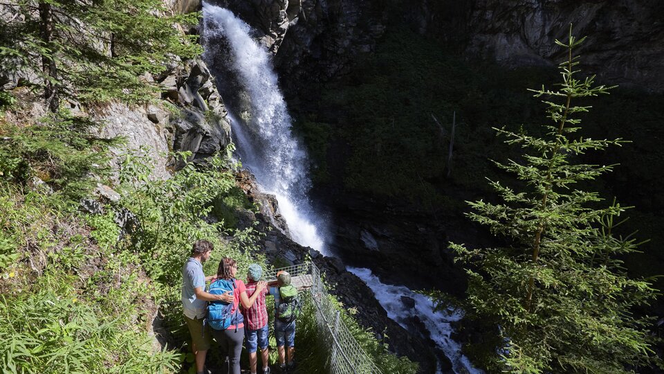 Eine Gruppe von Menschen steht an einem Wasserfall in der Natur. Umgeben von Bäumen und üppiger Vegetation genießen sie die Aussicht.