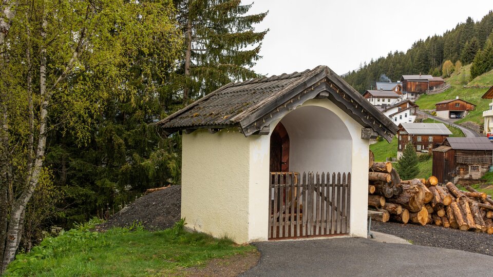 Eine kleine, charmante Hütte mit einem abgewinkelten Dach steht an einem Weg. Im Hintergrund sind Holzstapel und einige Häuser in einer bewaldeten Berglandschaft zu sehen. | © Bernhard Gruber – Photography