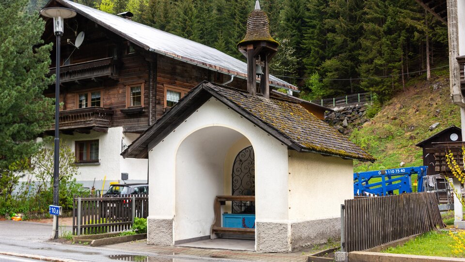 Eine kleine, weiße Kapelle mit einem Dachreiter steht an der Straße. Im Hintergrund sind typische alpine Holzhäuser und Bäume zu sehen. | © Bernhard Gruber – Photography