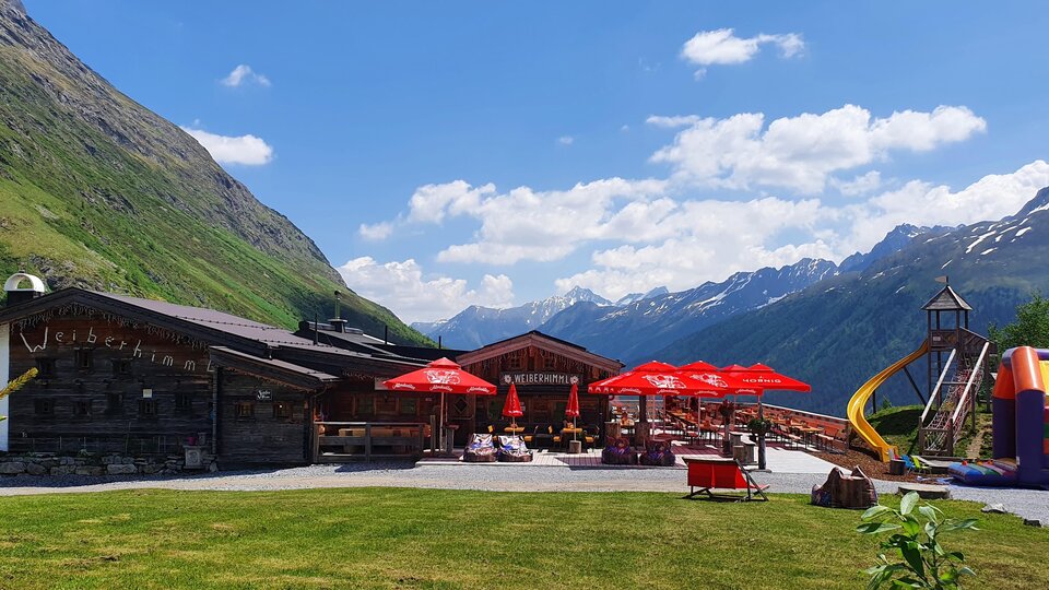 Ein gemütliches Gasthaus in den Bergen mit roten Sonnenschirmen. Im Hintergrund sind grüne Wiesen und majestätische Berge zu sehen.