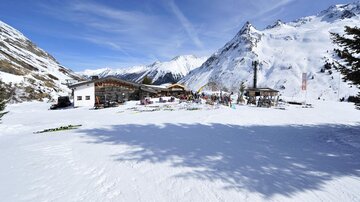Eine malerische Berglandschaft bei Schnee mit einem kleinen Gebäude und Menschen, die sich im Freien aufhalten. Im Hintergrund sind beeindruckende Berge sichtbar.