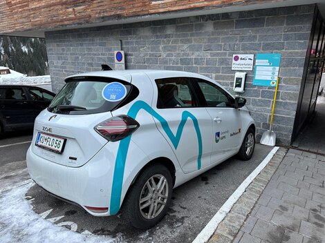 An electric car is parked at a charging station in a snow-covered environment. It is located at the edge of a parking lot, surrounded by gray stone facade.