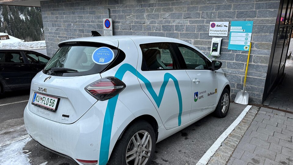 An electric car is parked at a charging station in a snow-covered environment. It is located at the edge of a parking lot, surrounded by gray stone facade.